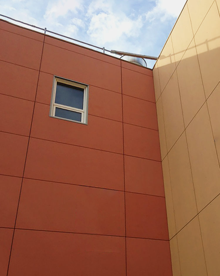 Modern building corner with intersecting red and yellow facades, featuring clean lines, a single rectangular window, and visible rooftop railing under a partly cloudy sky.