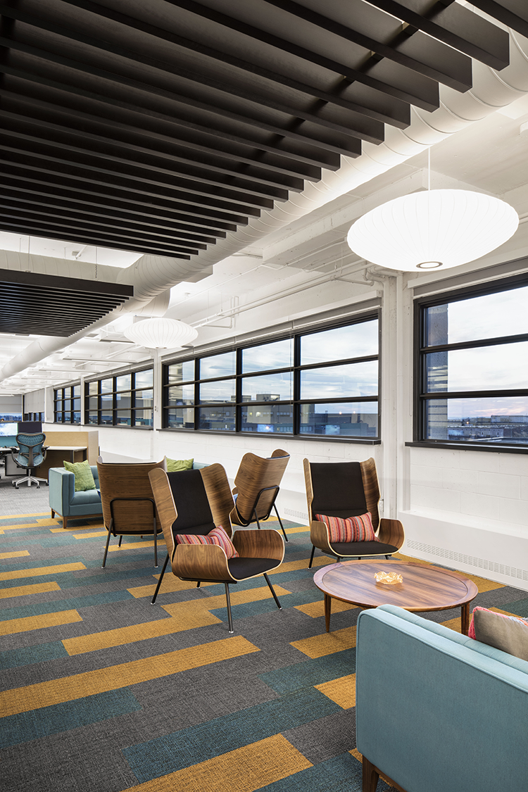 A stylish lounge space with modern armchairs around circular tables. The floor is covered with colorful patterned carpet tiles in blue, green, yellow, and gray. The ceiling has black slatted panels and round pendant lights. Large windows let in natural light and offer outdoor views.