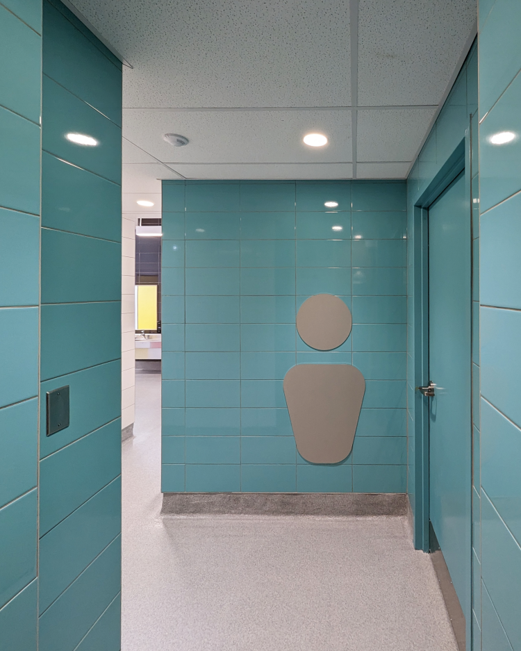 A hallway with teal-colored tiled walls and a stylized symbol representing a boy, indicating a boys' restroom. A door is visible to the right, and the space is brightly lit. The floor is light grey, and the ceiling has recessed lighting. White tiled walls can be seen further inside the space.