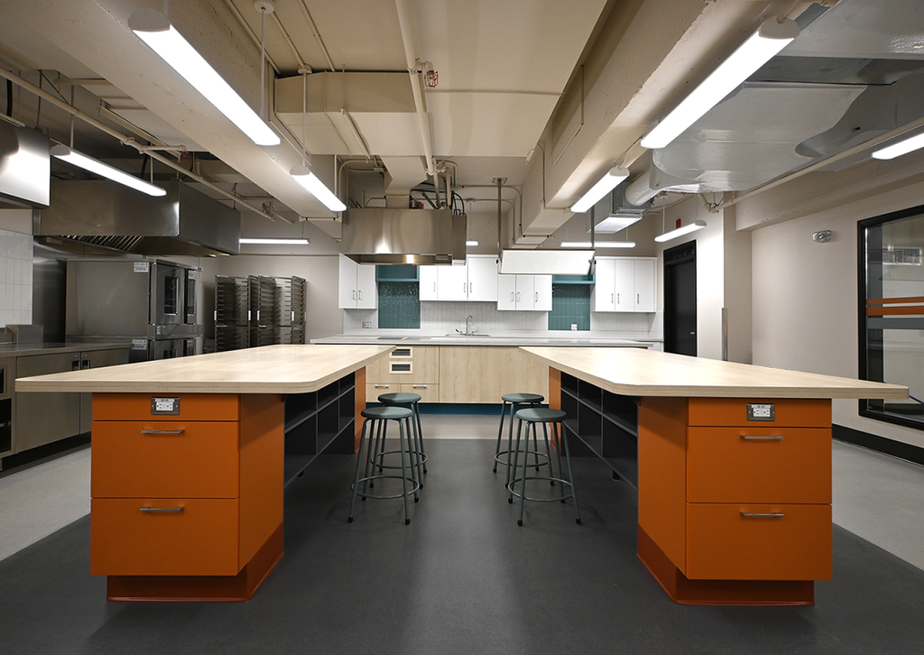 A modern teaching kitchen with two large workstations that have orange bases and open shelving. Each workstation has metal stools, and the kitchen features stainless steel appliances, industrial lighting, and cabinetry with wood and white finishes. Overhead are visible pipes and vents, adding to the industrial aesthetic.