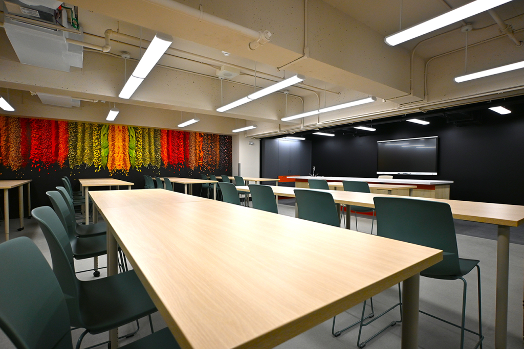 A brightly lit classroom with light wood tables and teal chairs arranged in rows. A large colorful mural of cascading spices in red, orange, yellow, and green decorates the back wall. At the front, there is a teaching station with a digital screen mounted on a black wall.