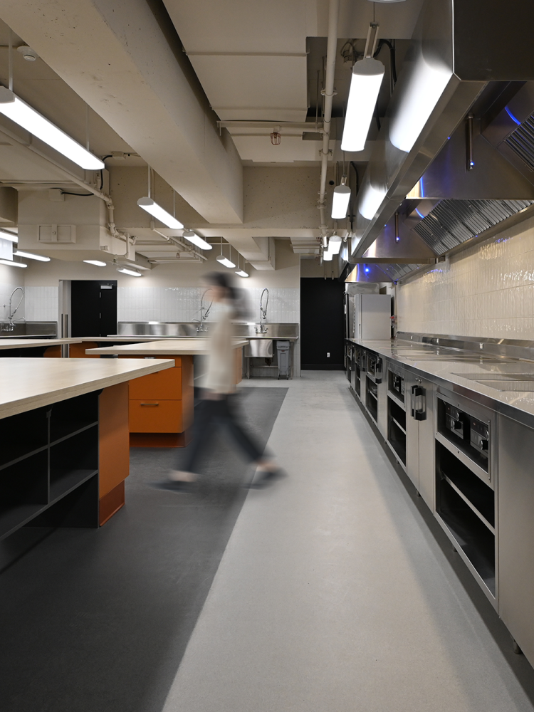 A side view of the teaching kitchen showing a person walking past stainless steel cooking stations with built-in stoves and ventilation hoods. The orange workstations and overhead lighting continue the industrial theme. Sinks and metal racks are visible in the background.