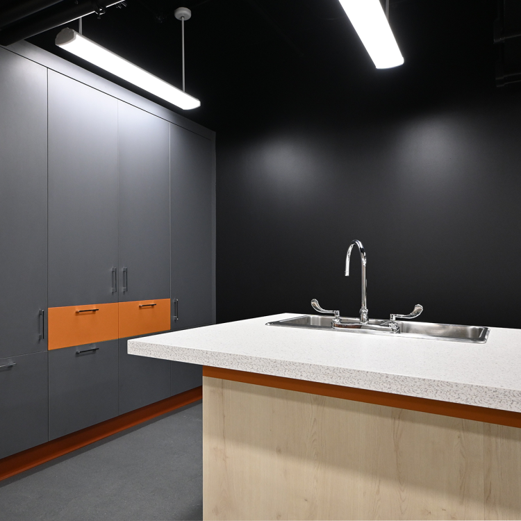 Teaching kitchen featuring a central island with a light wood base and white countertop, integrated stainless steel sink, and wall-mounted matte grey cabinets accented with burnt orange drawers. Set against a black wall with industrial lighting above.