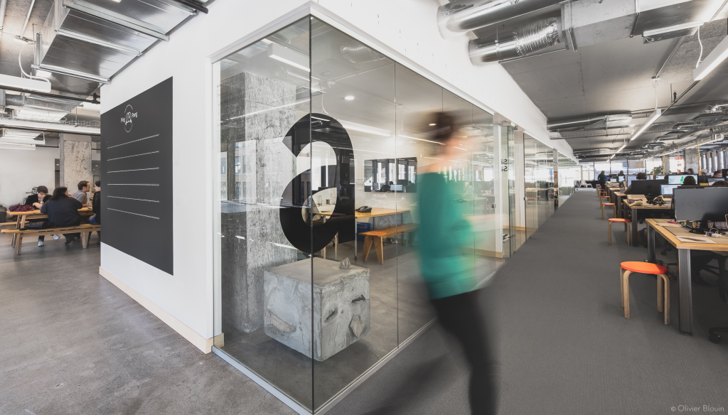 A corridor in a bustling office environment is in the background. Glass walls and a large black letter “a” displayed on the glass panels are in the foreground. Desks with computers line one side, indicating an active workspace. The floor is a polished concrete on the left side and carpet on the right. The ceiling consists of exposed ductwork and suspended lighting fixtures.
