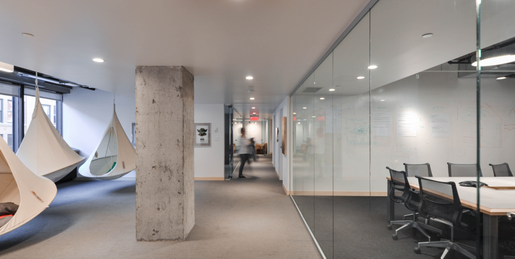 A modern office hallway features hammock-style hanging chairs on the left, suspended between large windows that allow natural light to fill the space and a central concrete pillar. The right side includes a glass-walled conference room with chairs arranged around a table. The floor concrete, except for the conference room which has carpeting.