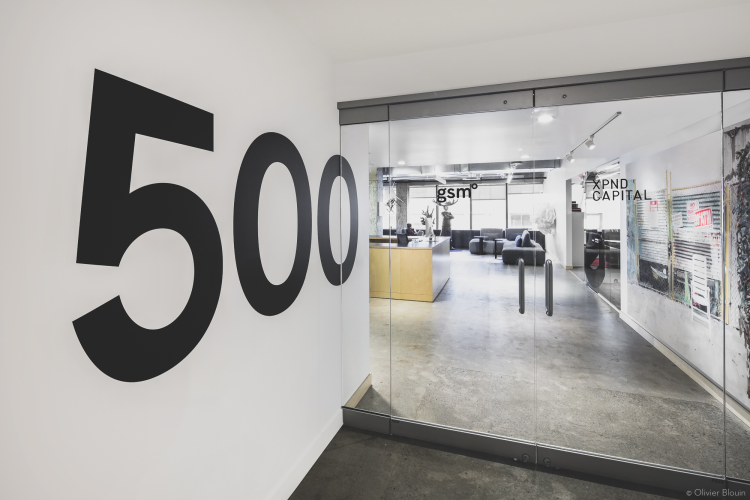 Entrance to a sleek office space marked by large black numbers “500” on a wall adjacent to glass doors. Through the doors, an open office area is visible with modern seating arrangements and signage for “gsm” and “XPND CAPITAL.”