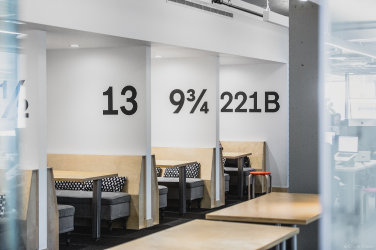 Office seating area with multiple small booths, each marked with unique identifiers such as “13,” “9¾,” and “221B,” in black bold letter. The booths feature cushioned bench seating and fixed tables. The floor is carpeted in a solid dark grey colour, and the ceiling has exposed piping and suspended lighting.