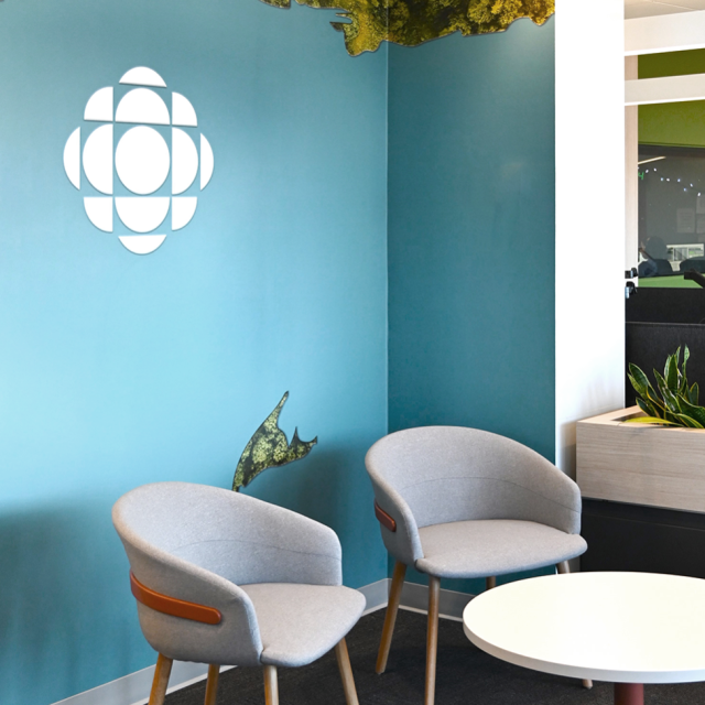 Contemporary lounge area featuring two grey upholstered chairs with wooden legs, a round white table, and a teal accent wall with a white Radio Canada logo. A forest mural runs along the top edge of the wall, adding a natural element to the space.