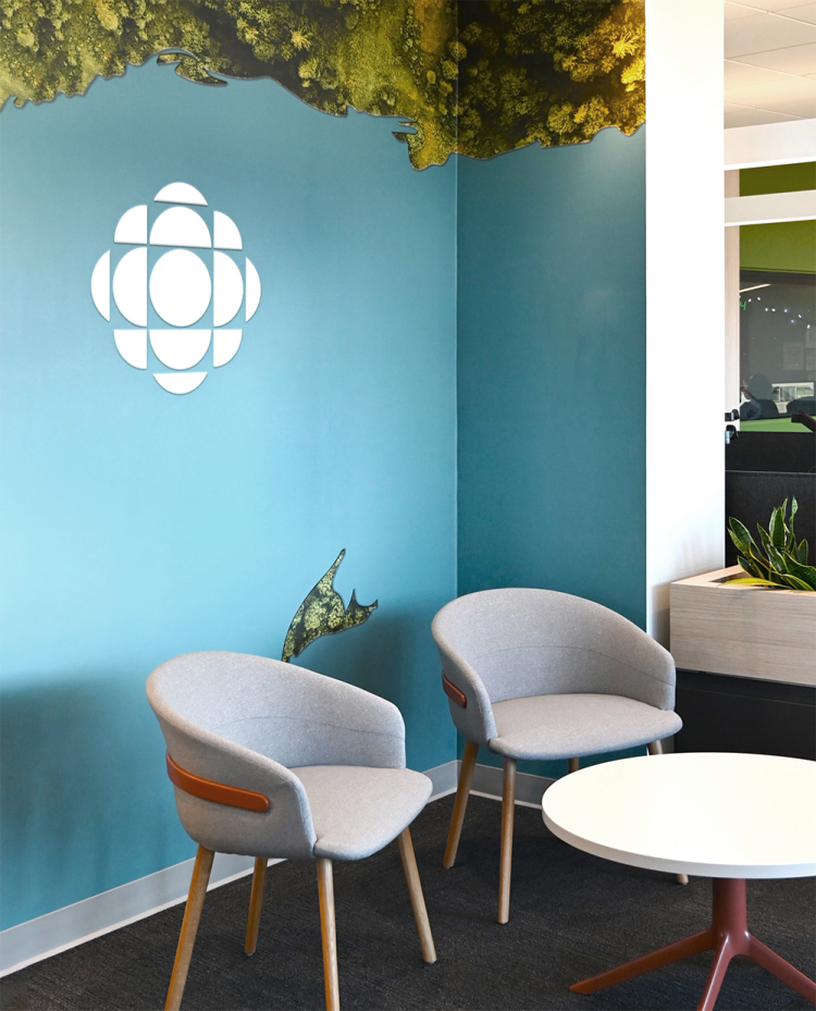 Contemporary lounge area featuring two grey upholstered chairs with wooden legs, a round white table, and a teal accent wall with a white Radio Canada logo. A forest mural runs along the top edge of the wall, adding a natural element to the space.
