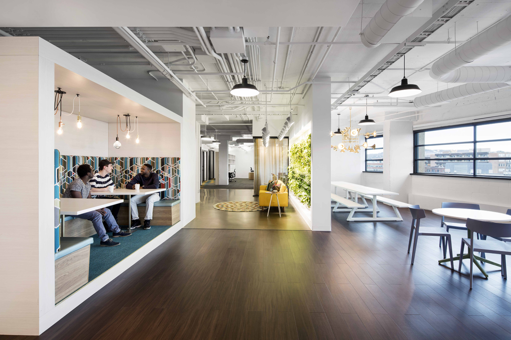 Contemporary office lounge with three men sitting and chatting in a booth with colorful geometric-patterned upholstery. A yellow couch faces a green plant wall in the center. The far end includes long white picnic-style tables near large windows with a city view.