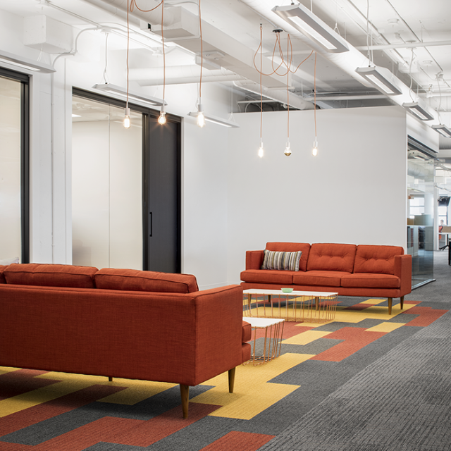 Contemporary office lounge area with mid-century modern red sofas, warm accent lighting, and a patterned carpet in shades of red, yellow, and gray.