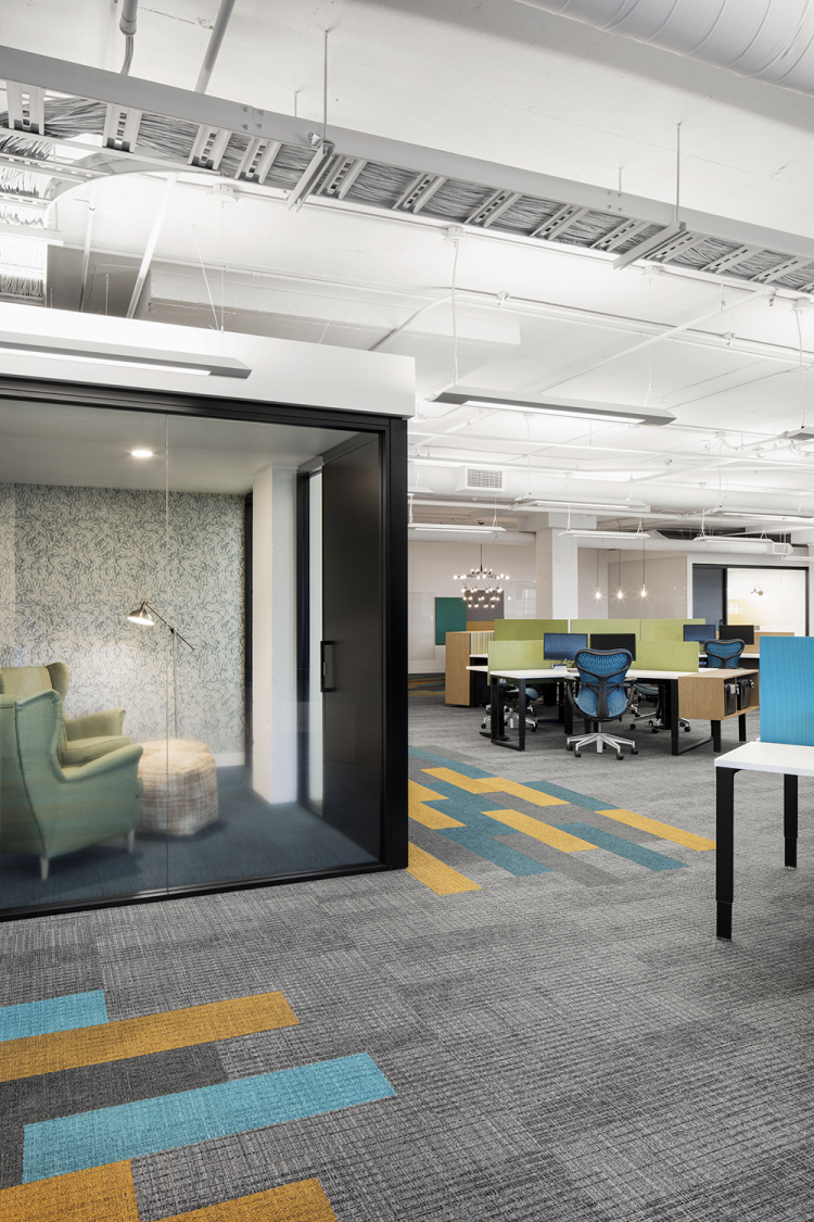An open office area with ergonomic chairs and partitioned desks arranged in rows. The floor is covered with patterned carpet tiles in gray, yellow, and blue. The ceiling is white with exposed ductwork and suspended rectangular light fixtures. To the side, a small enclosed lounge with soft seating and a floor lamp.