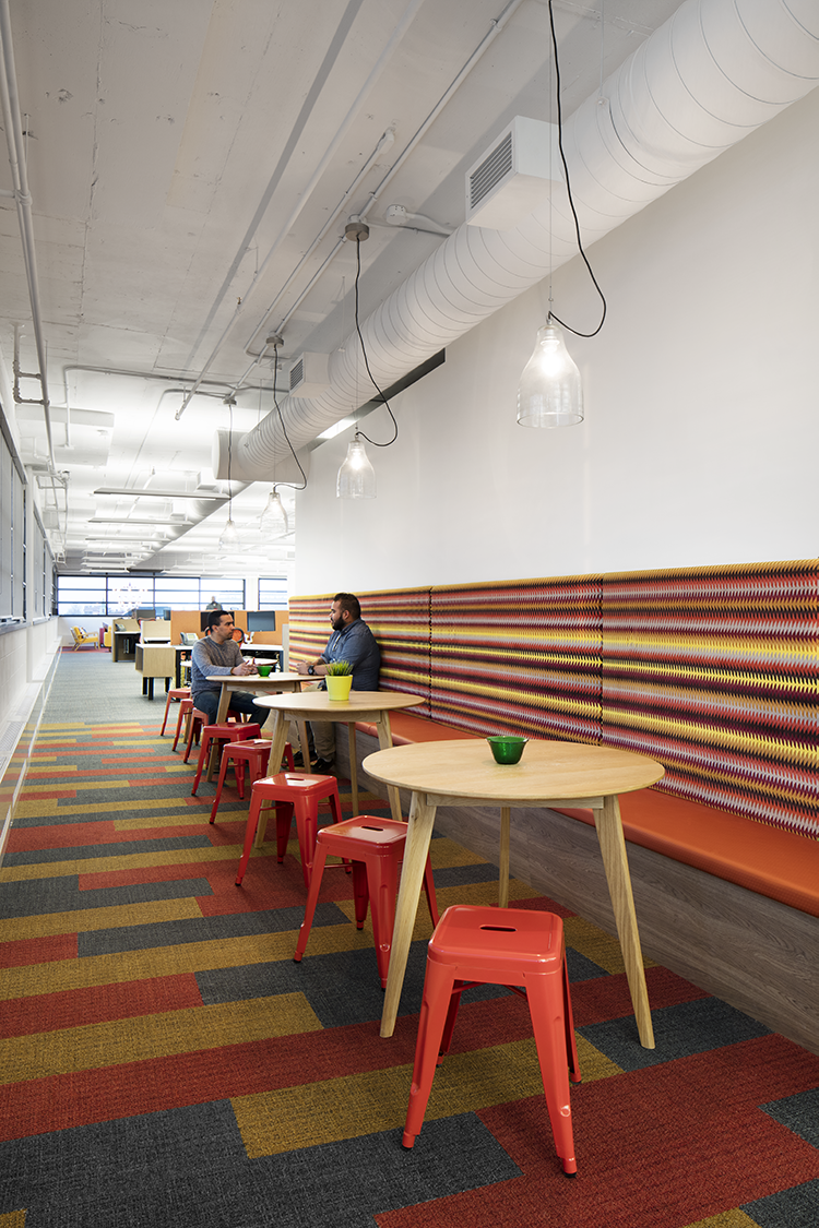 Two individuals sit at one of the three wooden round tables along a wall with a built-in bench of bold, colorful striped patterns. The flooring consists of vibrant carpet tiles in red, orange, and green. The ceiling is white with exposed ductwork and hanging pendant lights.