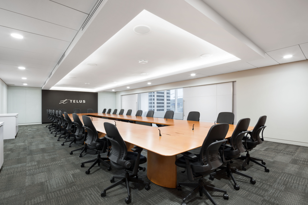 The image features the TELUS conference room, showcasing the long wooden meeting table surrounded by black office chairs. The room is illuminated by recessed ceiling lights andd diffused lighting. The “TELUS” logo is visible on a black wall,.