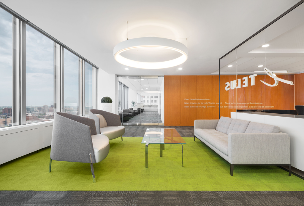 Modern office waiting area with tall windows, vibrant green carpet, contemporary grey and white seating, and a circular overhead light fixture. A glass partition seperates the space from the reception.