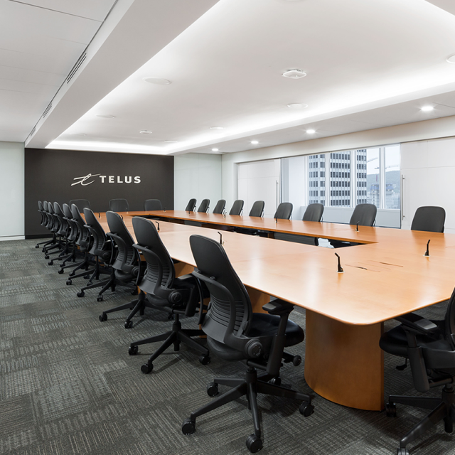 The image features the TELUS conference room, showcasing the long wooden meeting table surrounded by black office chairs. The room is illuminated by recessed ceiling lights andd diffused lighting. The “TELUS” logo is visible on a black wall,.