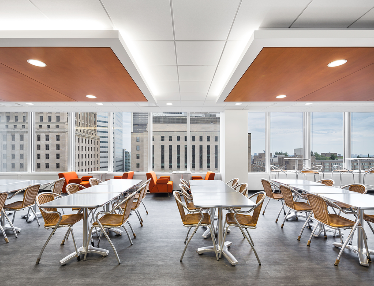 Bright office cafeteria with large windows offering city views, featuring metal-framed tables and rattan chairs. Warm wood ceiling panels and matching lounge seating add warm contrast to the modern, clean design.
