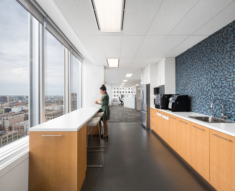 A modern office kitchen area with large windows offering expansive city views. The space features light wood cabinetry, white countertops, and a stainless steel sink. A long counter with white stools are placed by the window.
