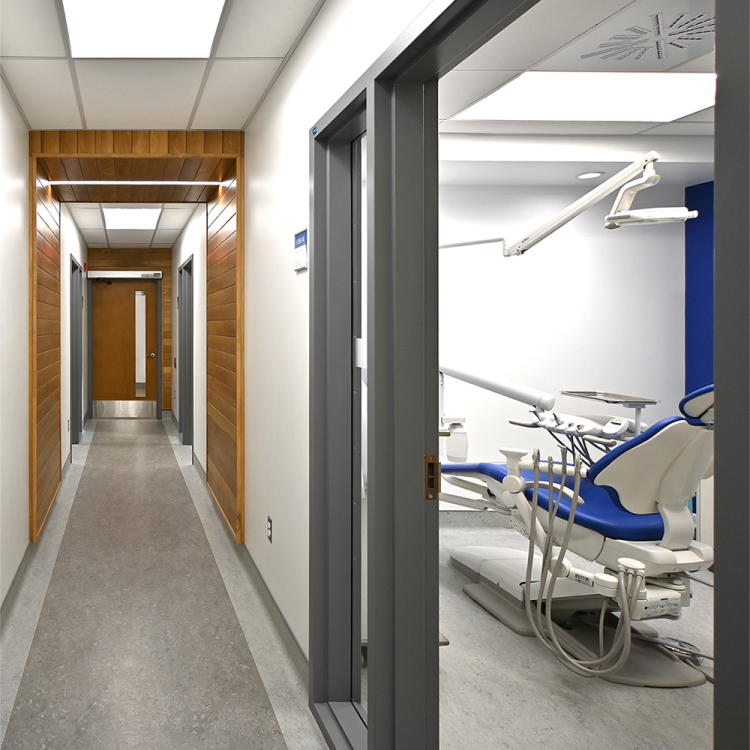 Clean, modern corridor in a dental clinic featuring light gray flooring, white walls, and warm wood paneling with recessed lighting overhead. Multiple treatment rooms are accessible from the hallway