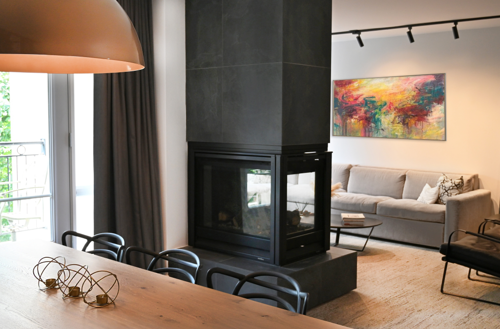 View of a modern hotel-room living area with a central three-sided fireplace, beige sectional sofa, black accent chairs, and a vibrant abstract painting on the wall, adjacent to a dining area with natural light from a large window.