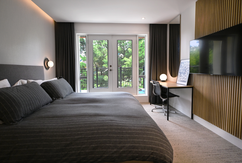 Wide view of a modern bedroom showcasing a large bed with dark pinstripe bedding, a sleek wall-mounted TV on wooden slats, and a minimalist workspace by full-length windows and doors that frame lush outdoor scenery.