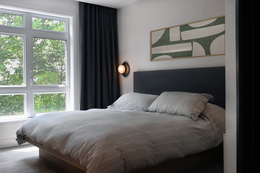 Contemporary bedroom featuring a low-profile wooden bed with a grey upholstered headboard, abstract wall art, dark curtains, and large windows bringing in natural light and views of greenery outside.
