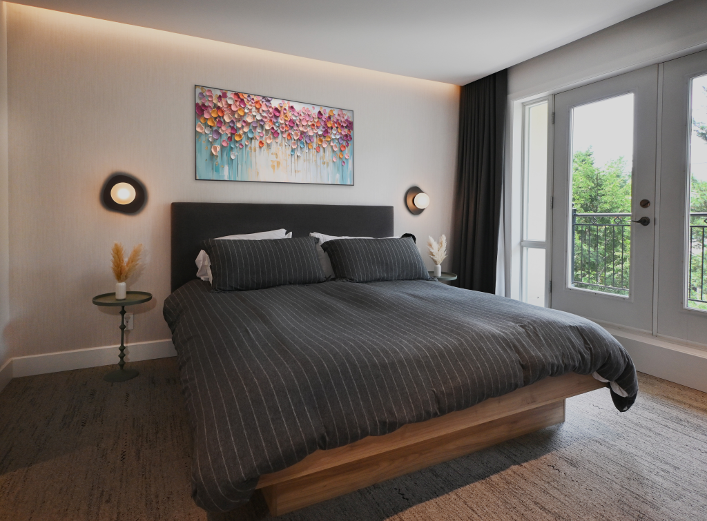 Elegant bedroom with a queen-size bed in charcoal grey bedding, colourful artwork above the headboard, warm ambient lighting, side tables with pampas grass, and doors opening to a balcony with lush outdoor views.