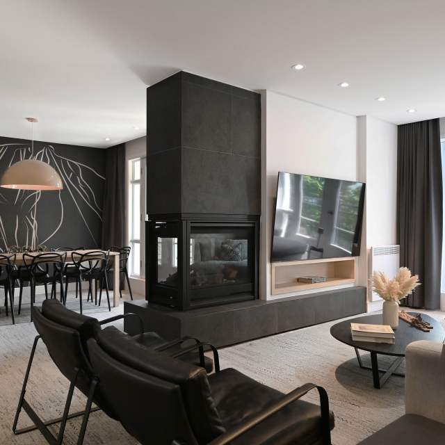 Contemporary open-concept hotel-room living and dining area featuring a dual-sided black stone fireplace, a wall-mounted TV, minimalist black chairs, and a large wall mural in the dining area with modern line art.
