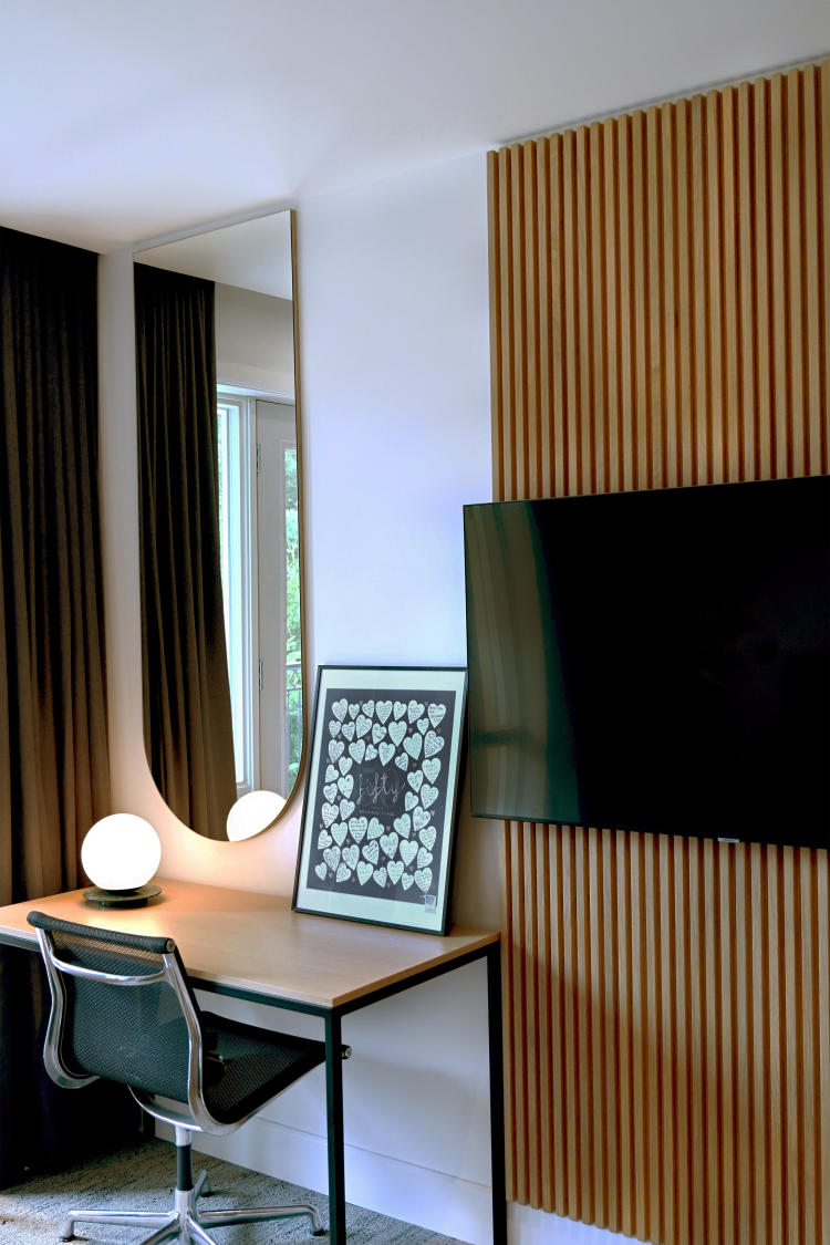 Modern work nook in a bedroom featuring a minimalist desk, mesh office chair, and a spherical table lamp beneath a tall arched mirror. A framed artwork rests against the wall beside a slatted wooden panel that integrates a wall-mounted flat-screen TV.