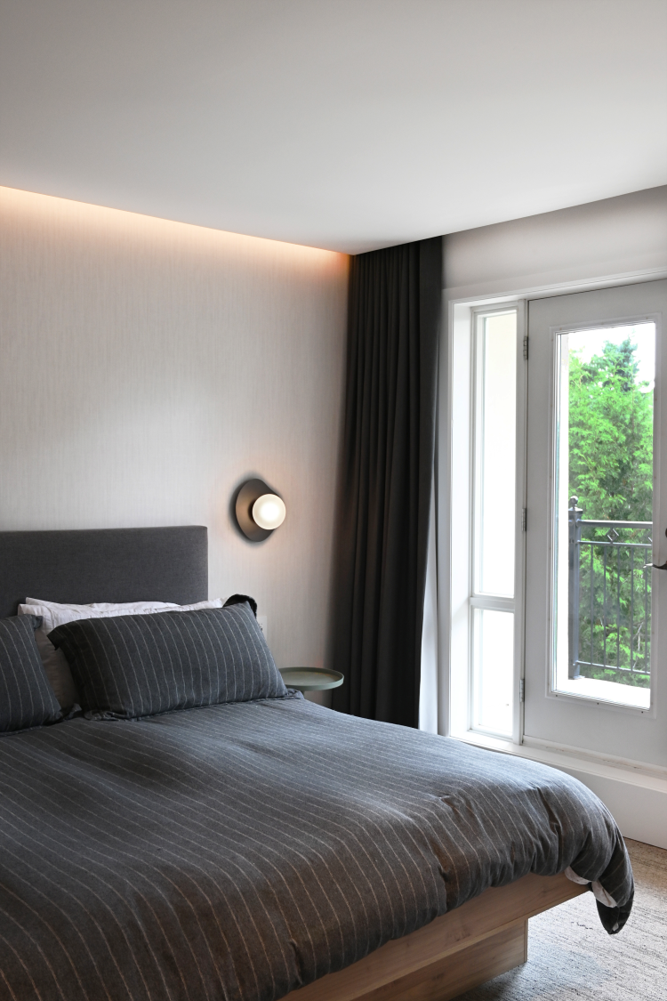 Contemporary bedroom with a charcoal upholstered headboard and matching bedding, complemented by soft wall lighting and full-height blackout curtains. Doors lead to a private balcony with views of greenery.
